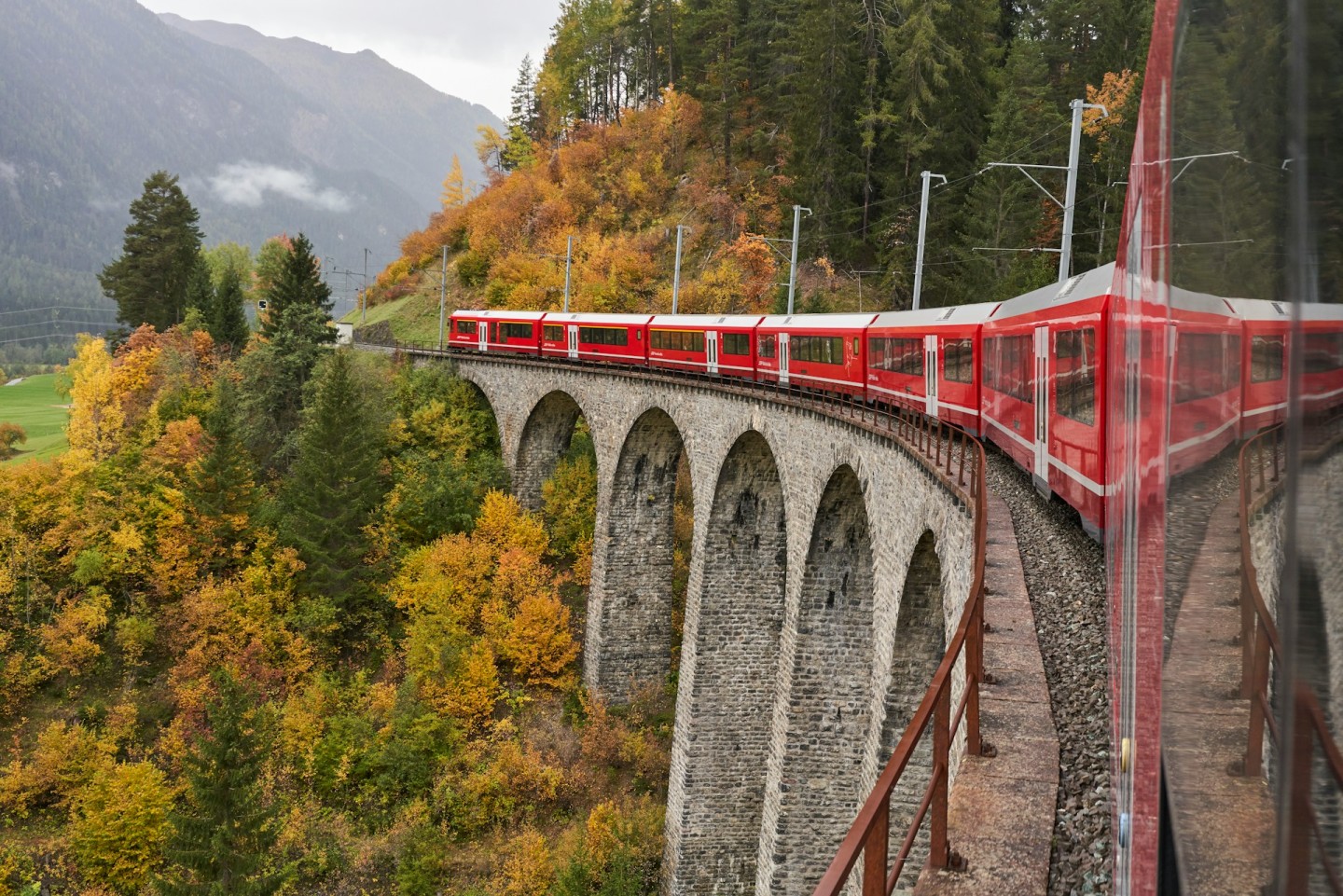 Red Rhaetian Railway train crossing a stone viaduct in the wooded Albula Valley.